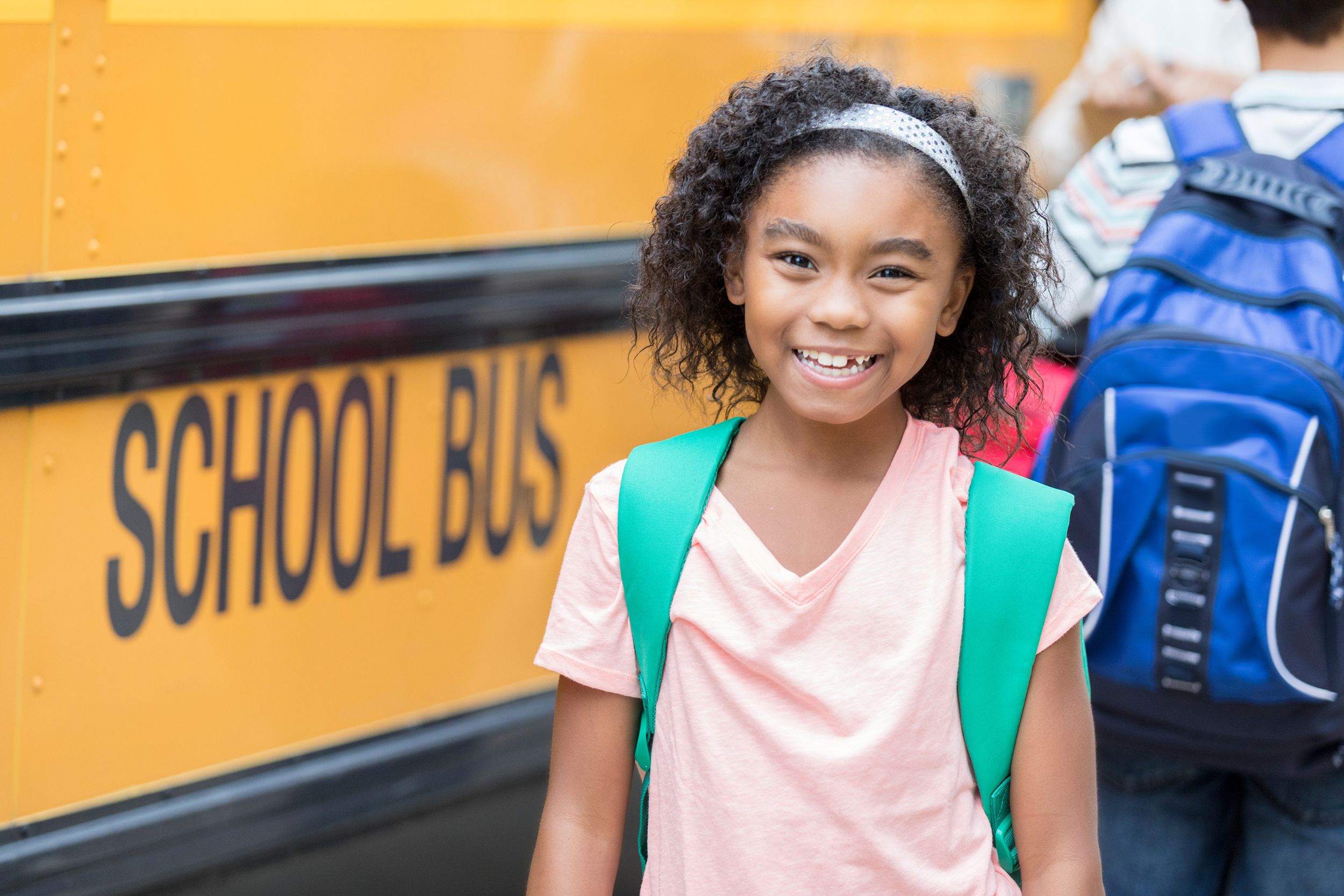 African American schoolgirl smiles for camera as classmates board bus