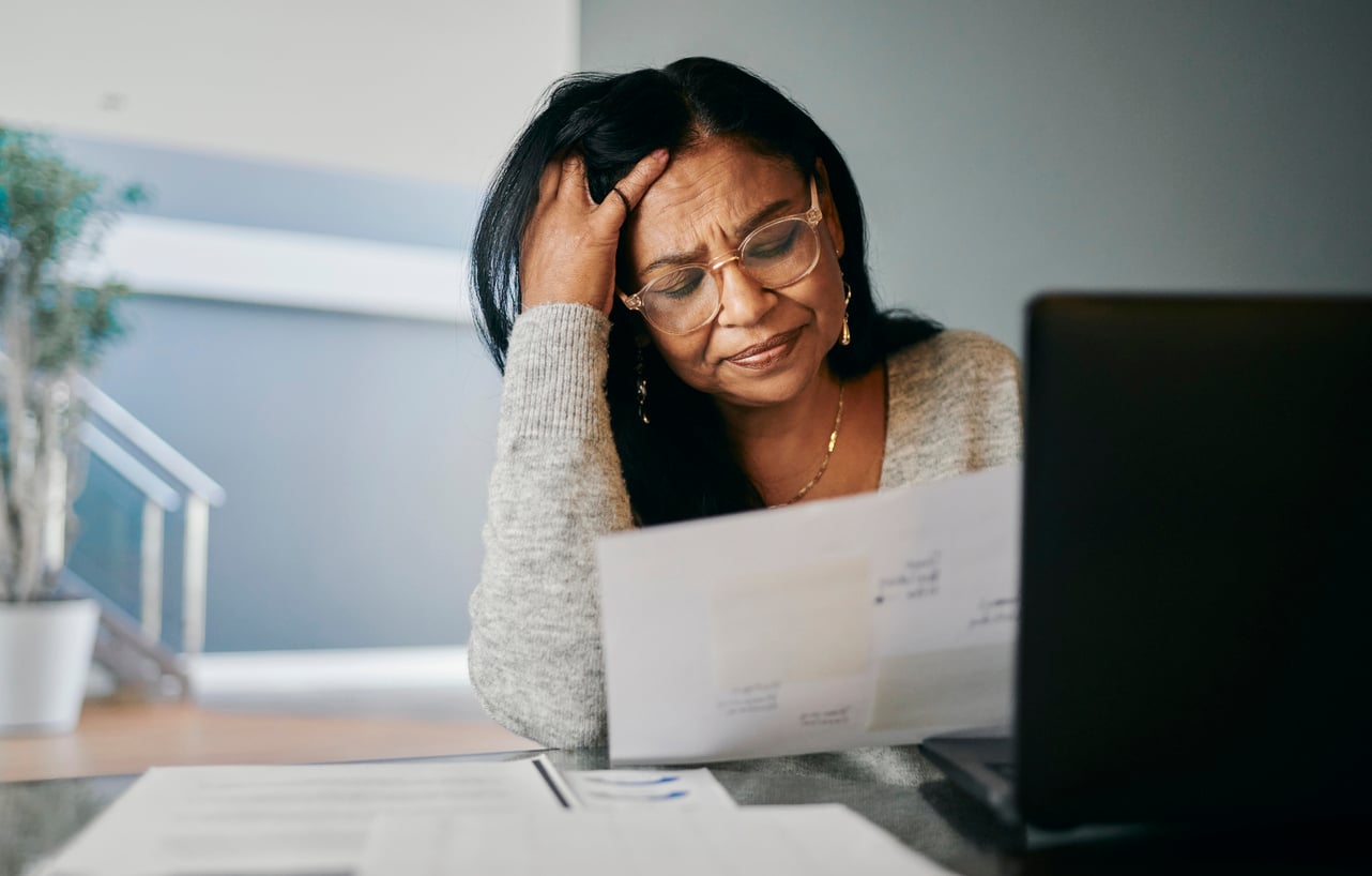 A woman sits at a table, looking stressed as she reads a document. She wears glasses, rests her head on one hand, and sits in front of an open laptop with more papers nearby.