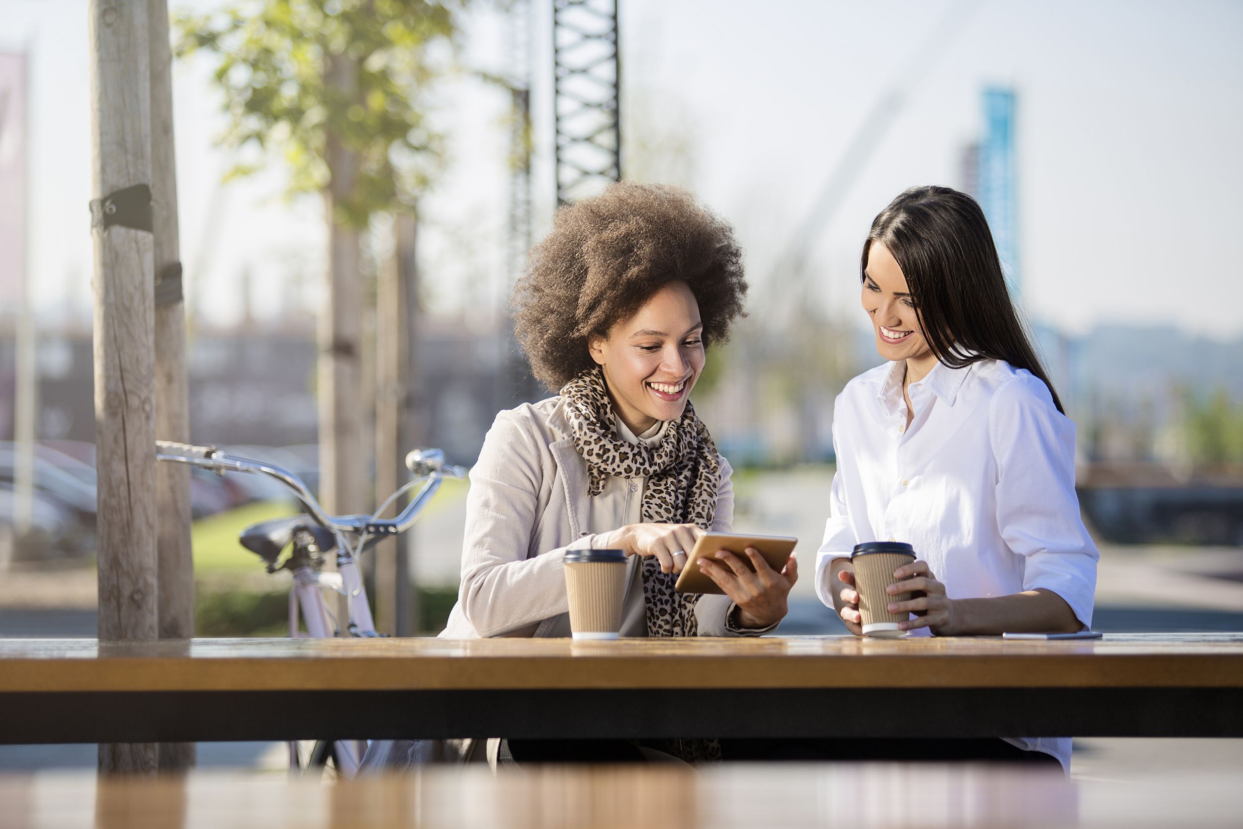 two women drinking coffee outside while sitting and looking at a tablet