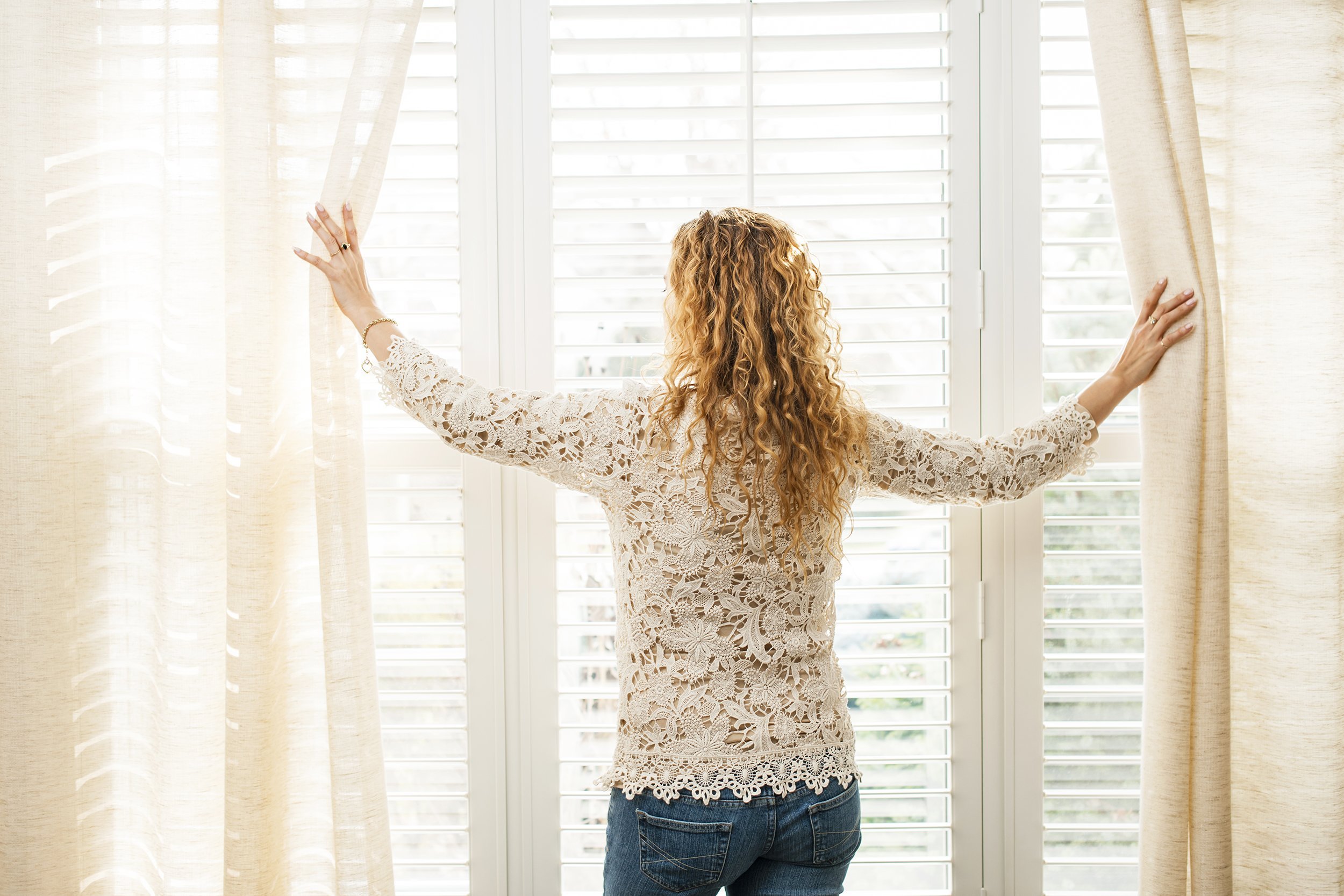 woman looking out big bright window with curtains and blinds