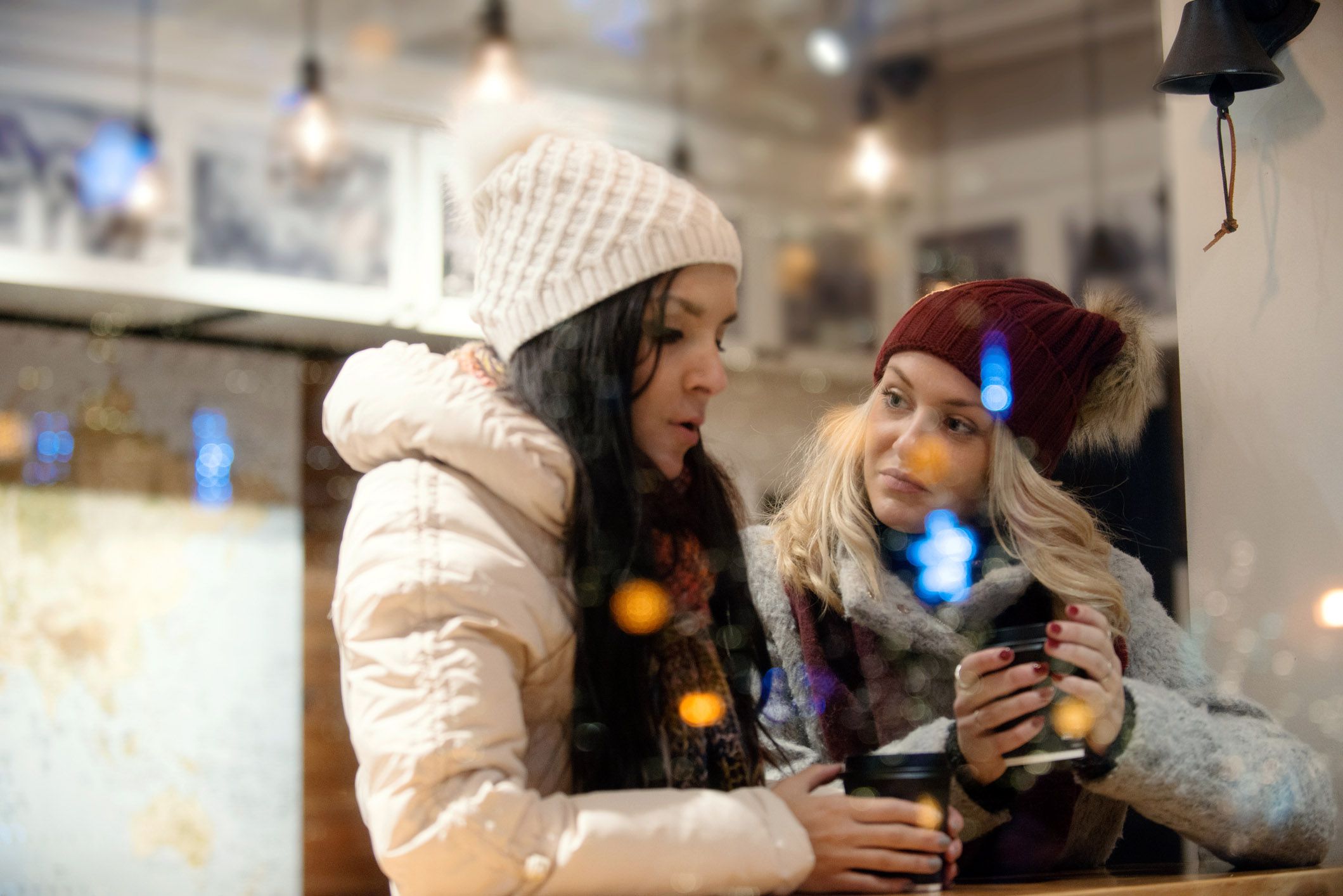 two friends in winter coats with christmas lights in background holding mugs having serious conversation