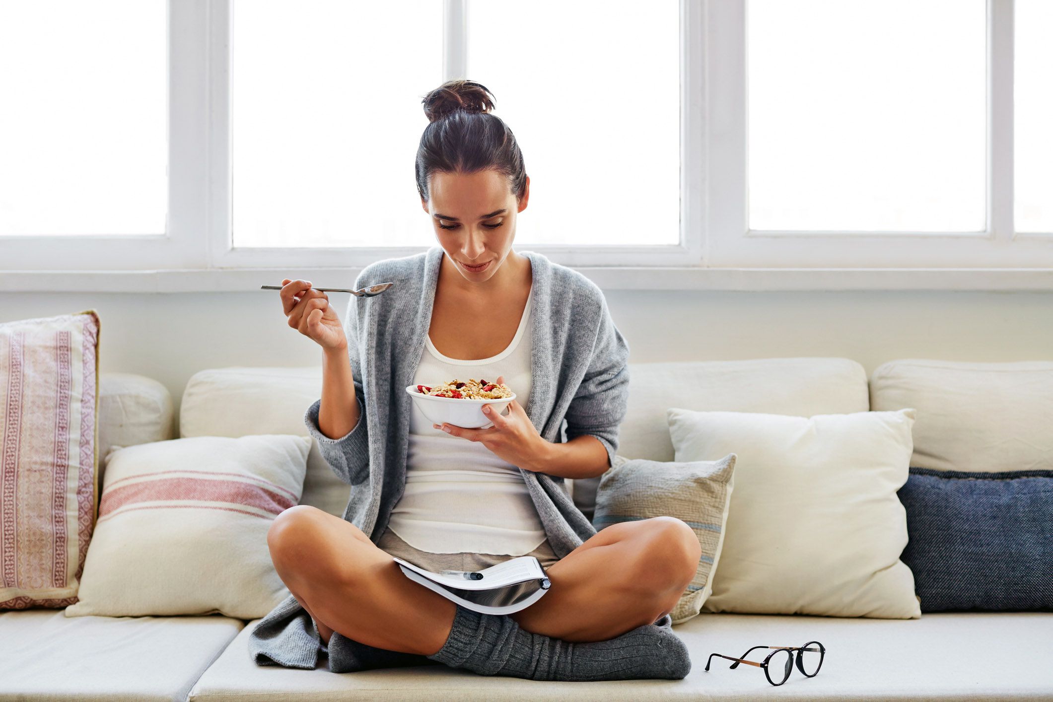 woman in sweater and cozy socks eating cereal with notebook and pen in lap