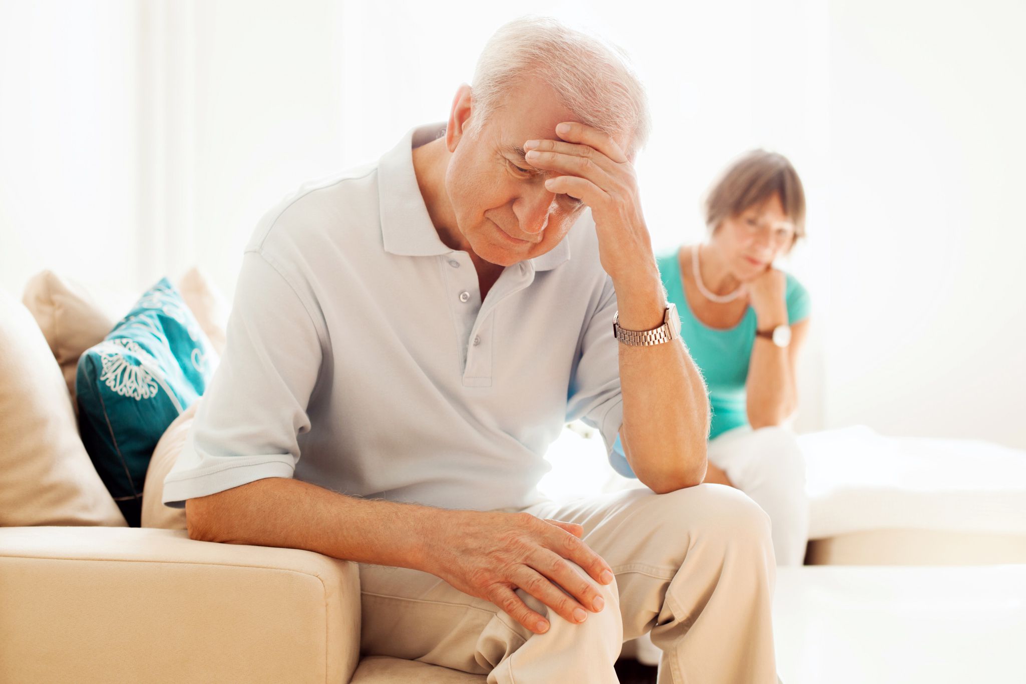 Close up of distressed senior man on couch holding his head with sad senior woman in the background on couch, after having an argument, sunlight shining into living room