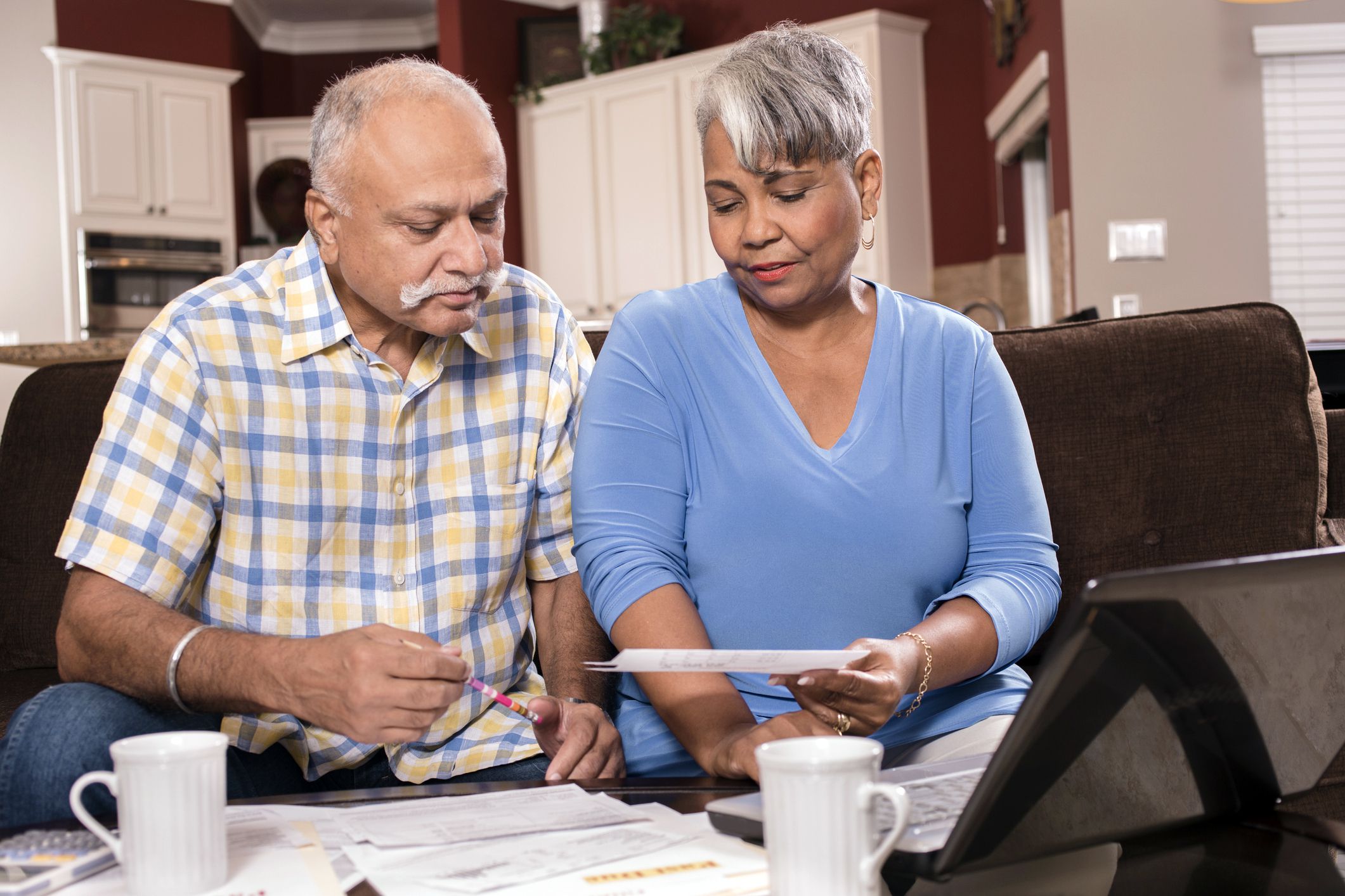 Senior couple sitting on couch looking over financial paperwork together while at laptop in the living room, with kitchen in the background