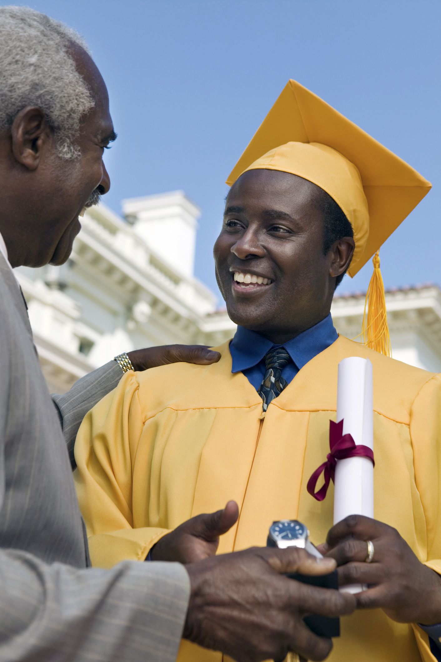 Senior father is congratulating son who just received his diploma during graduation, son is wearing gold graduation gown, father is in suit, college and blue sky in the background
