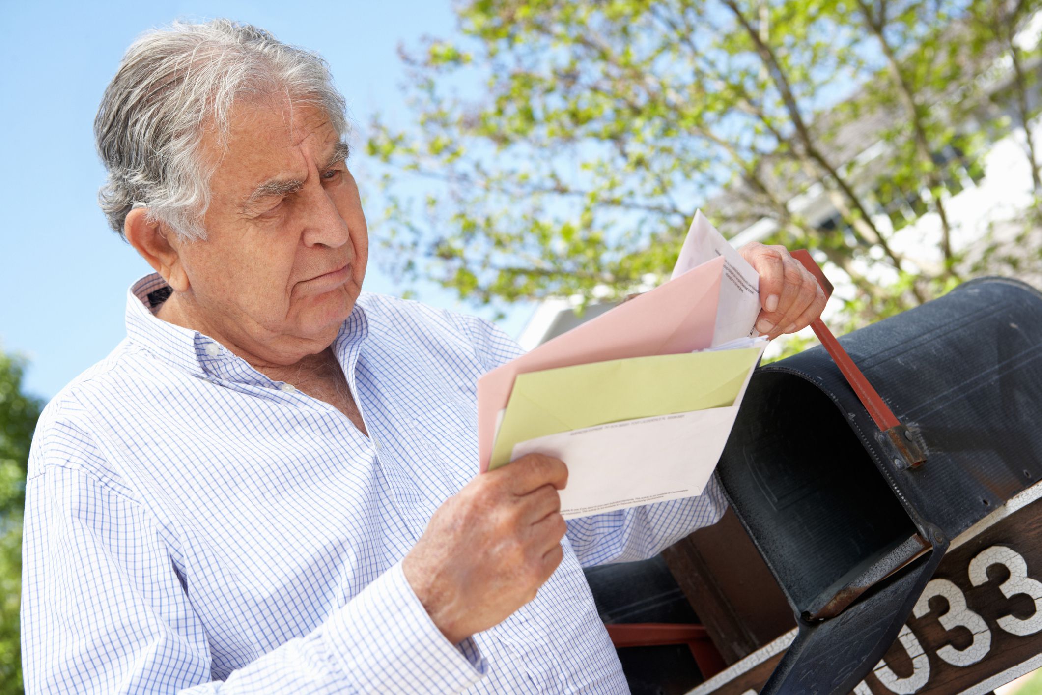 Senior man looking through mail while leaning on open mailbox, sunny spring day