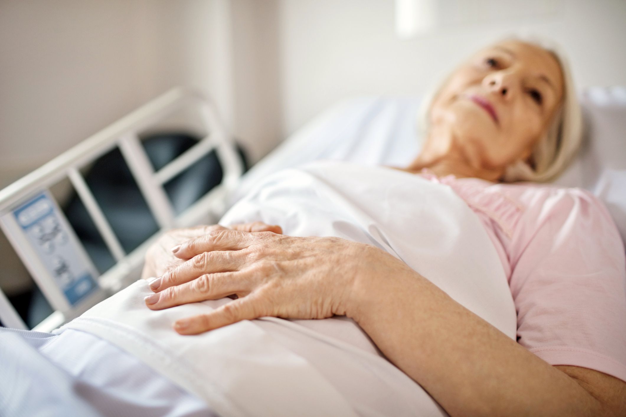 Senior woman awake laying face up in hospital bed, focus on hands together, blurred background of hospital room