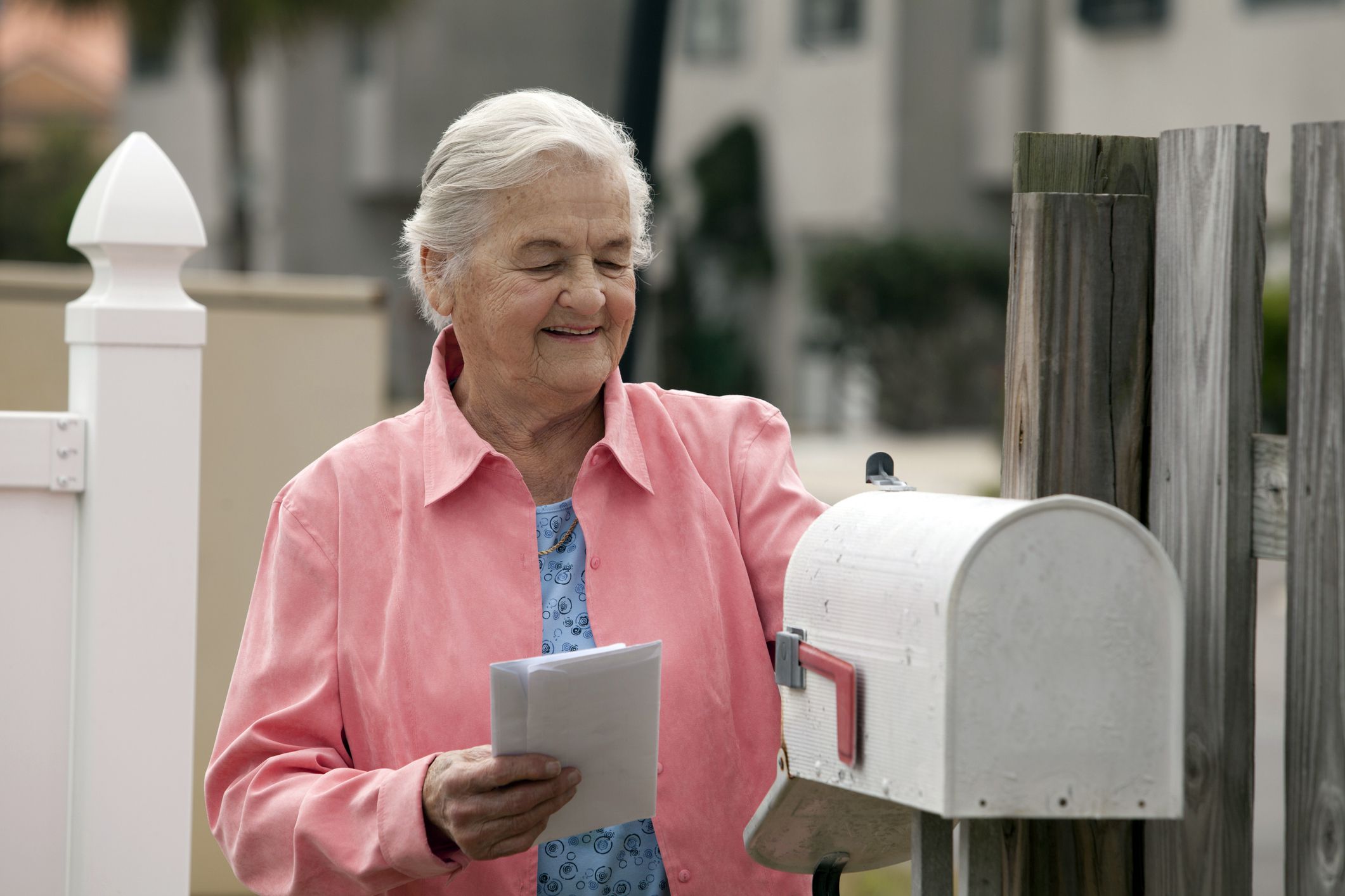 Smiling senior woman checking mail with mailbox Opened and she is holding two envelopes, fence then blurred apartments in background
