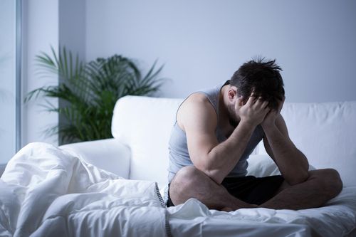 A man sits on the edge of a bed with his elbows on his knees and his head in his hands, appearing stressed or upset. The room is dimly lit with a white sofa and a plant in the background.
