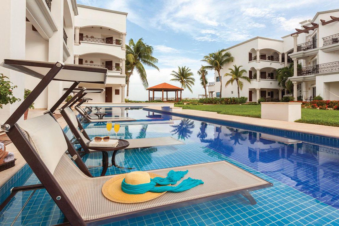 hat, scarf, glasses, book, mimosas on table and lawn chairs over pool, palm trees and suites in background