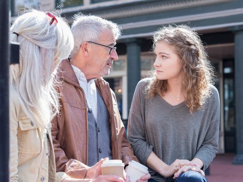 Three people, two older adults and a young woman, sit outside holding coffee cups. The older man and young woman are engaged in a serious conversation, while the older woman listens. They appear to be on a city street.