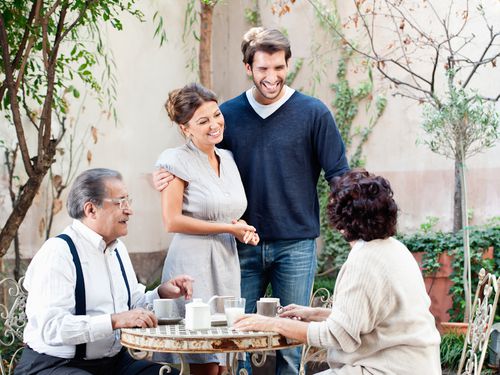 Two older adults sit at a garden table with coffee mugs, while a younger couple stands nearby smiling, enjoying a warm and cheerful outdoor gathering. Trees and plants surround the group.