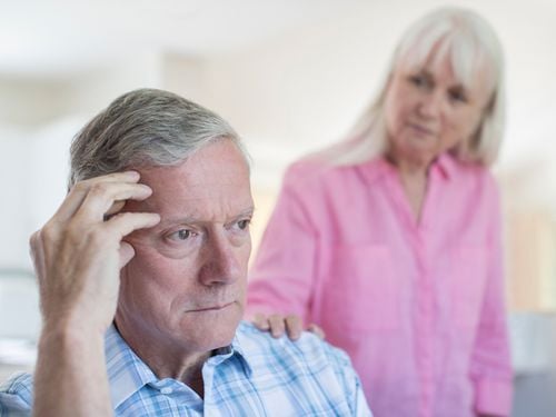 An older man sits looking worried or thoughtful, with his hand on his temple, while an older woman stands beside him with a concerned expression, resting her hand on his shoulder.