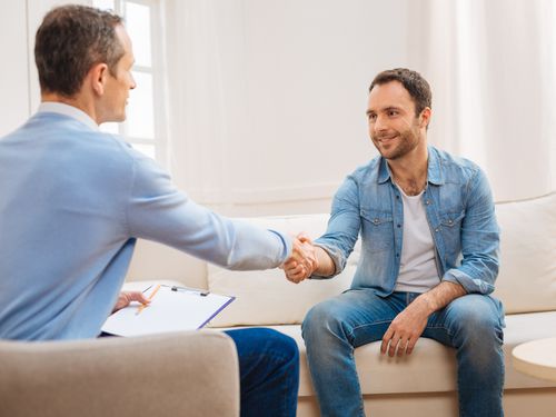 Two men sit facing each other on a couch in a bright room, shaking hands. One man holds a clipboard and pen, suggesting a professional or counseling setting.