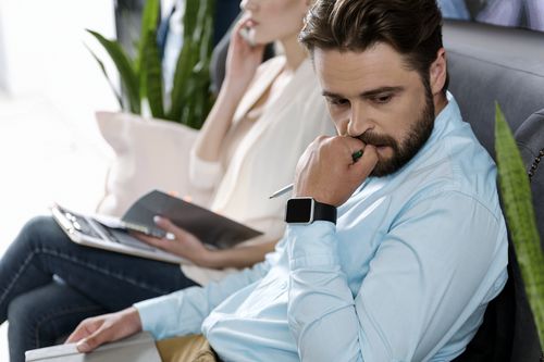 A man in a blue shirt sits pensively, holding a pen to his mouth, while a woman in the background works on a laptop and talks on the phone. Both appear thoughtful and focused.