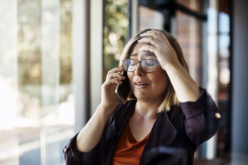 A woman with glasses looks distressed while talking on her phone, standing indoors by large windows with her hand on her forehead.
