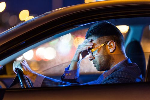 A man in glasses sits in a car at night, holding the steering wheel with one hand and resting his head on his other hand, looking stressed or tired. City lights blur in the background.