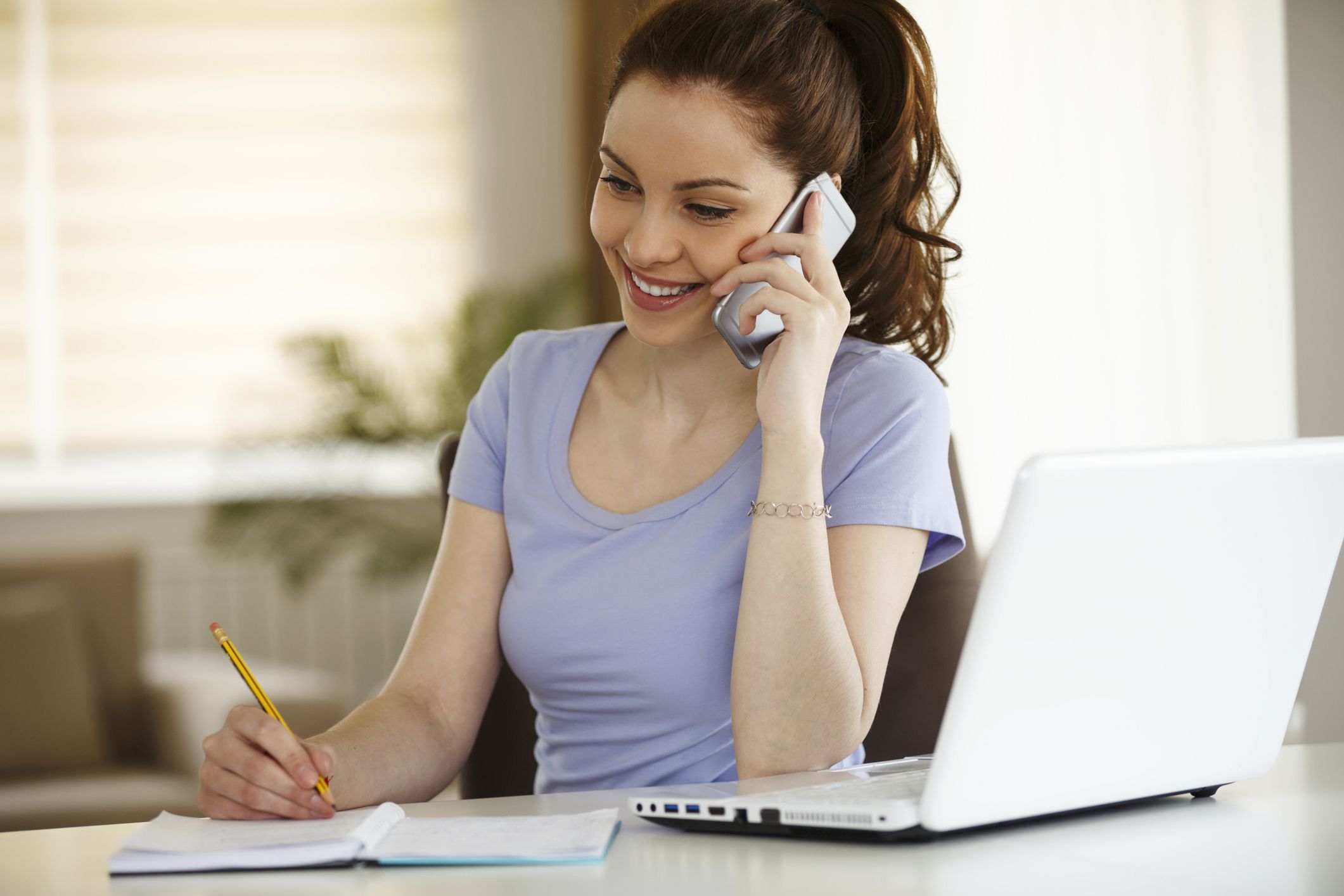 woman talking on phone with schedule open, pen in hand, laptop open on table