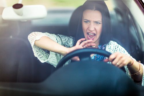 A woman sits in the driver’s seat of a car, looking distressed and yelling, with one hand pressed on the steering wheel, as if honking the horn.