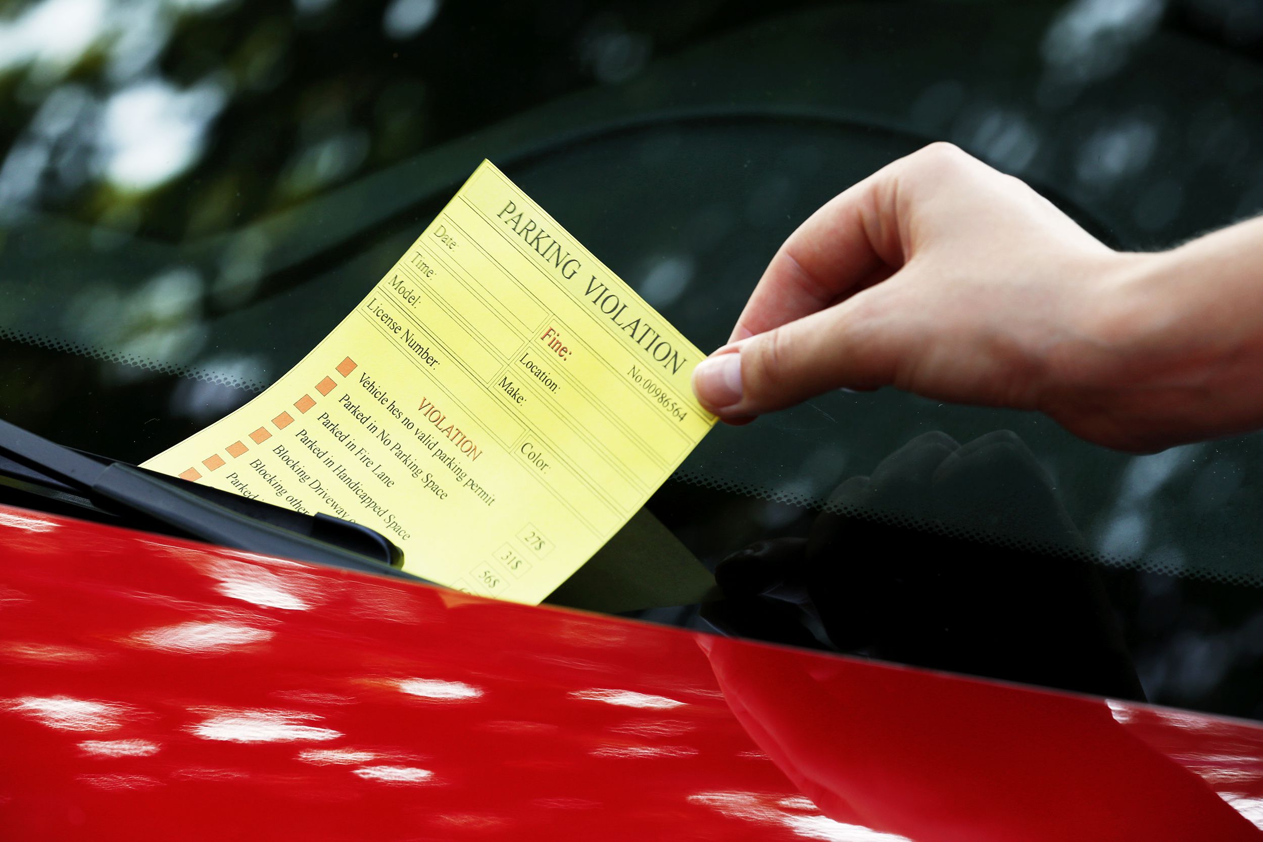 Hand picking up yellow parking violation ticket on the windscreen of a red car
