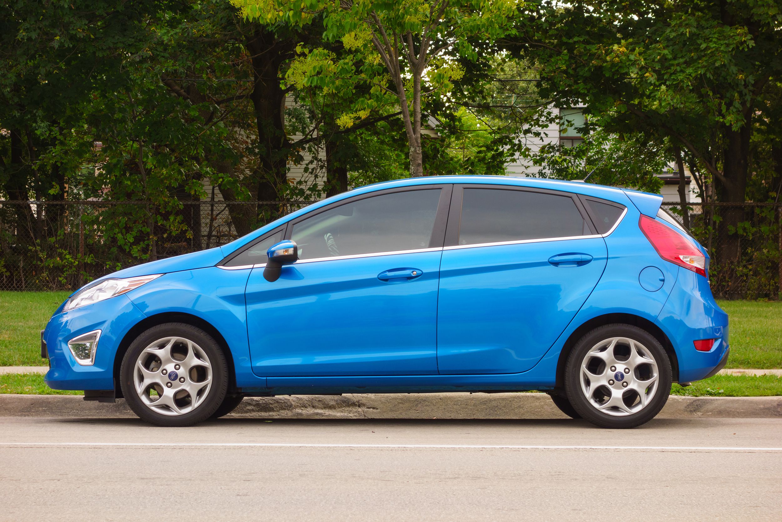 Left side-view of a blue colored Ford Fiesta car parked on the street, grass and trees in the background