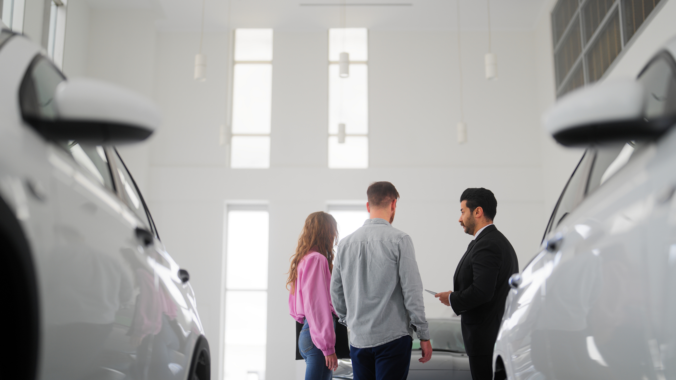 Rental car agent talking with couple, two white cars in the foreground, large white walls with windows