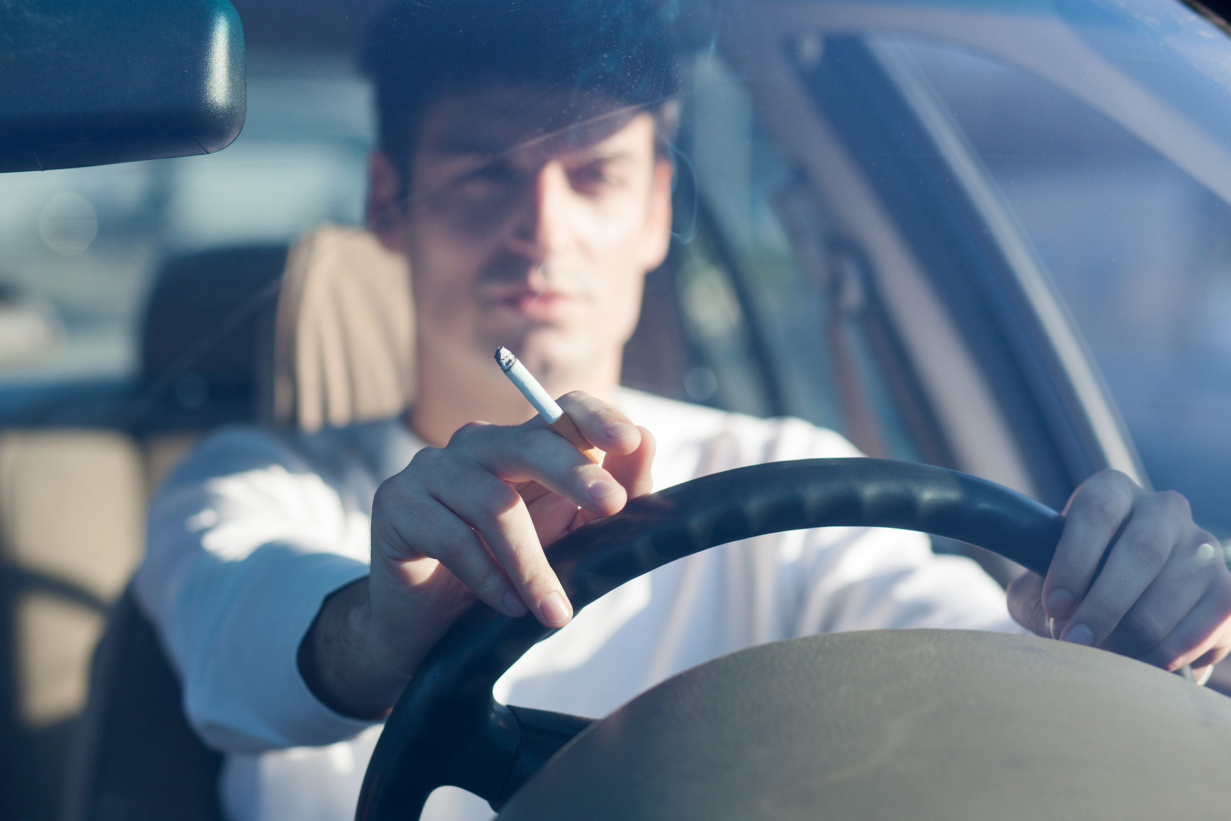 Smoking man driving with both hands on wheel, cigarette in fingers of right hand, selective focus, blurred background of inside of car