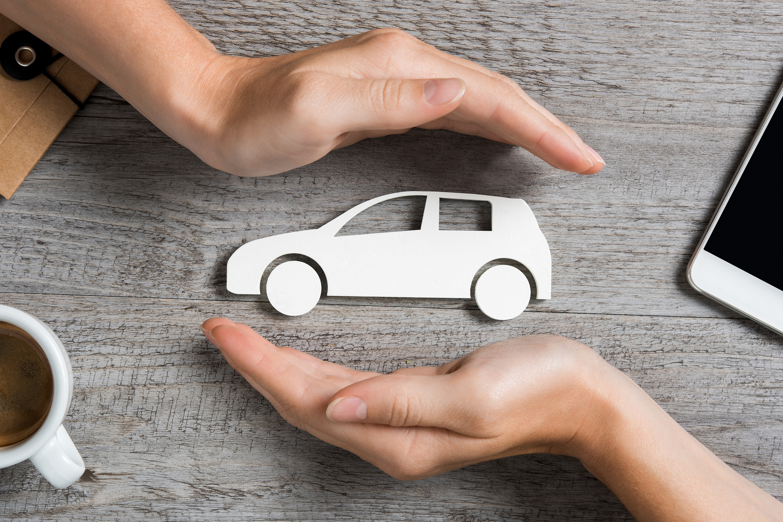 White paper car covered by two hands in a circle on the side on a grey table, cup of coffee, on left and table on right in the sides