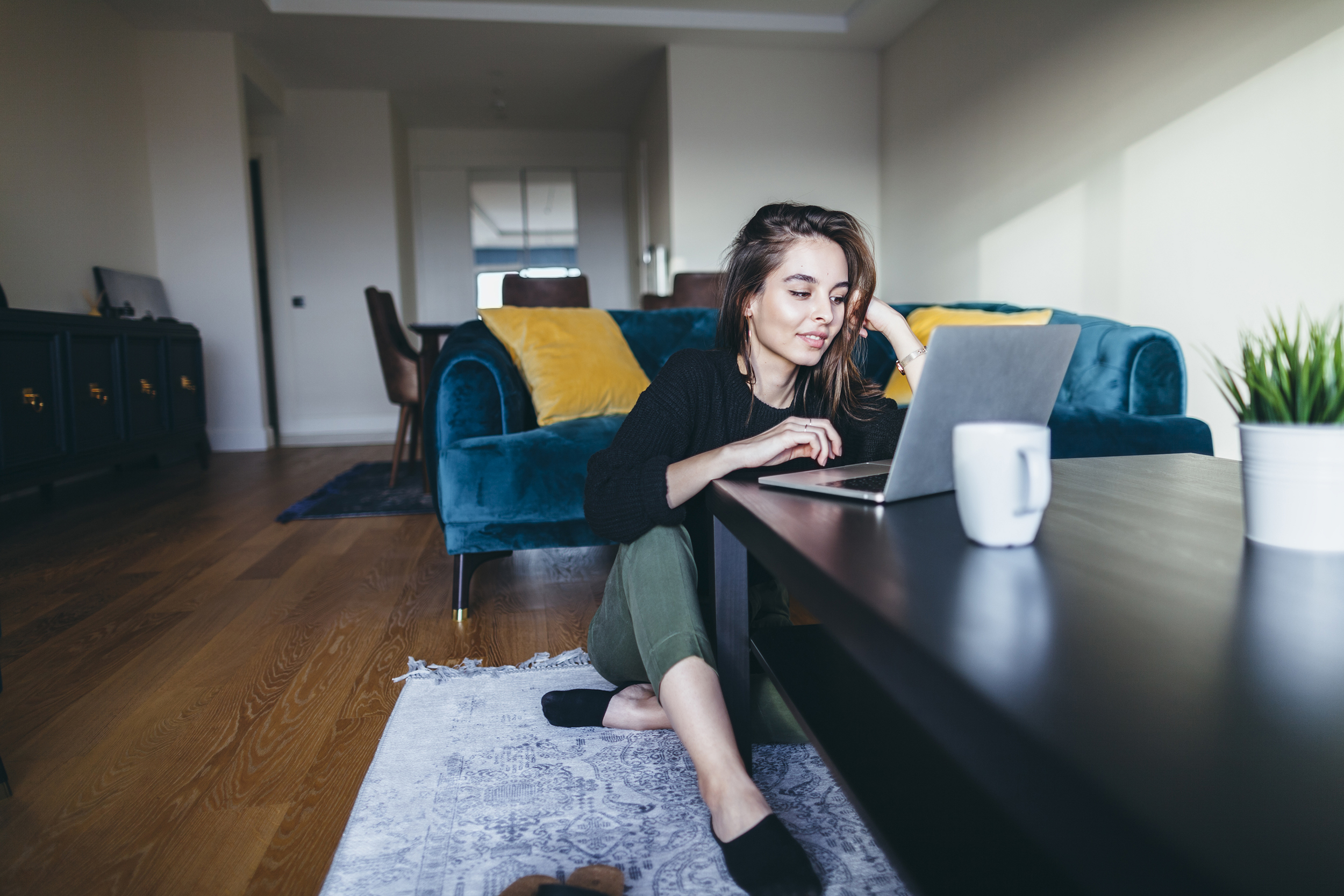 Woman sitting on the floor, using her laptop on a coffee table, in her living room, white walls of room in the background