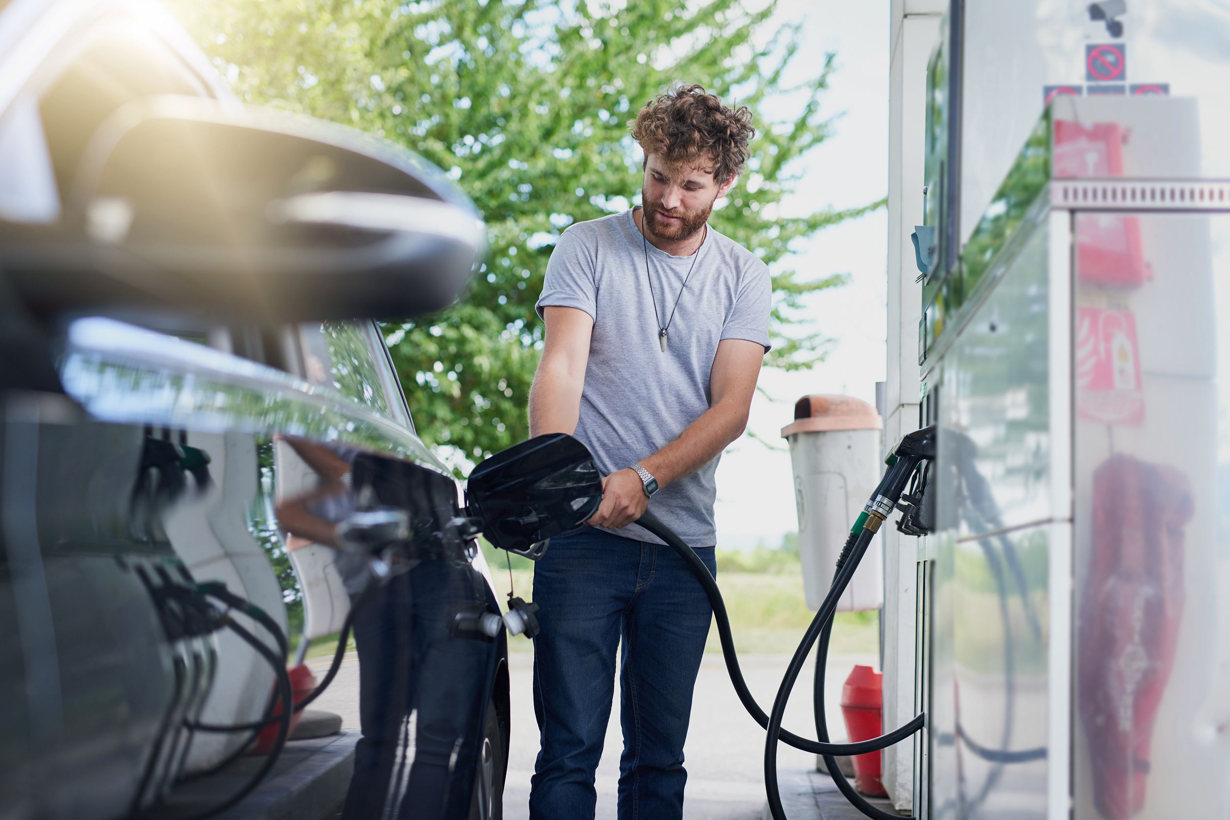 Young man using a gas pump to fill his car with gas, car on the left foreground, man in the middle, tree slightly blurred in the background
