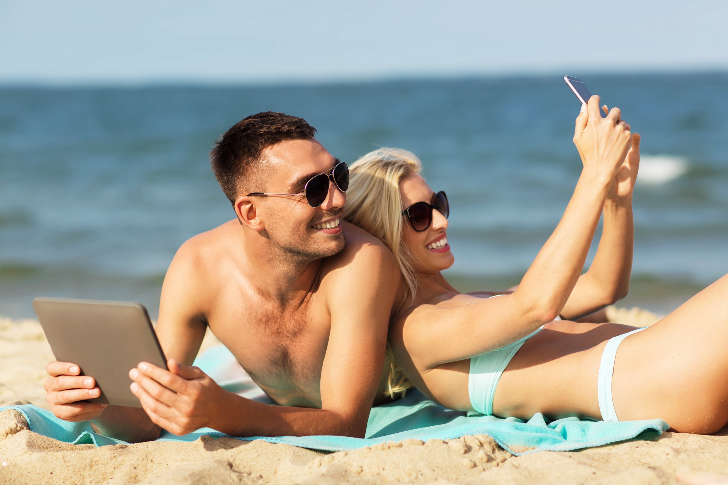 happy couple on vacation with tablet smartphone sunbathing on summer beach