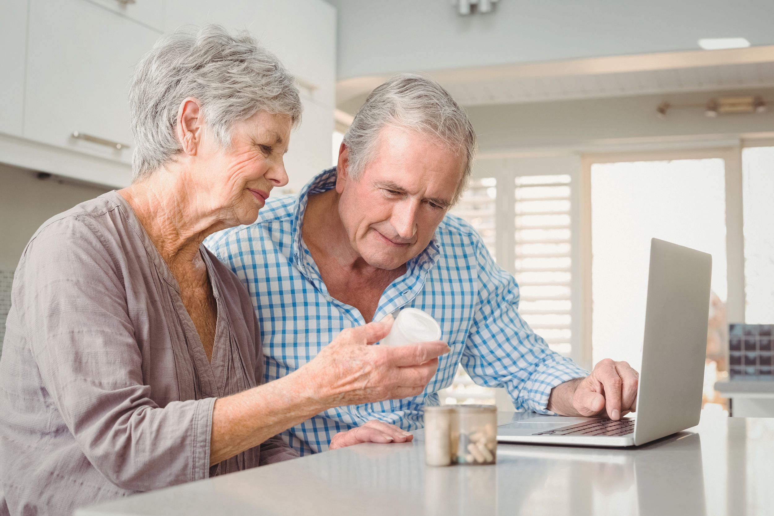 senior woman showing pills to husband with laptop in kitchen