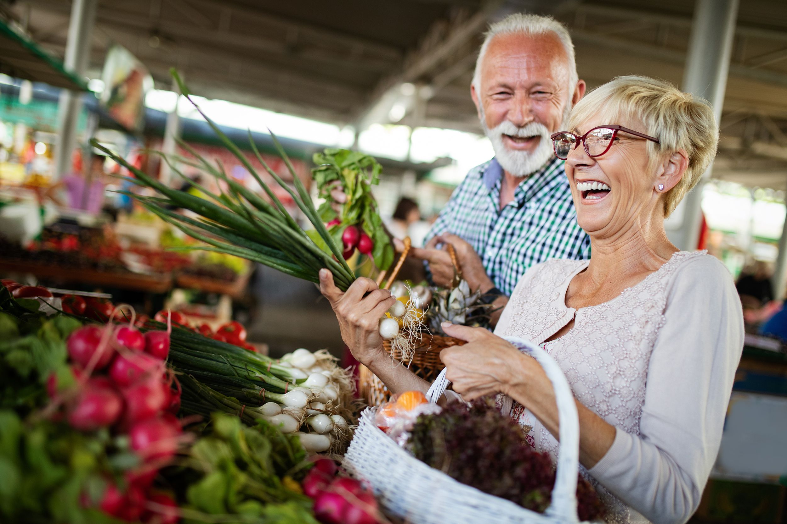 smiling senior couple holding basket with vegetables at the market