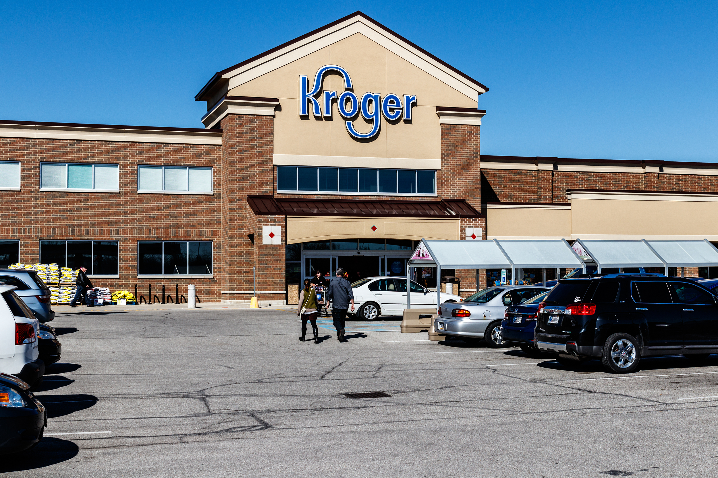 Front exterior of a Kroger grocery store, Indianapolis, parking lot in the foreground with cars and a few people walking towards the store, on a sunny day with a clear blue sky in the background