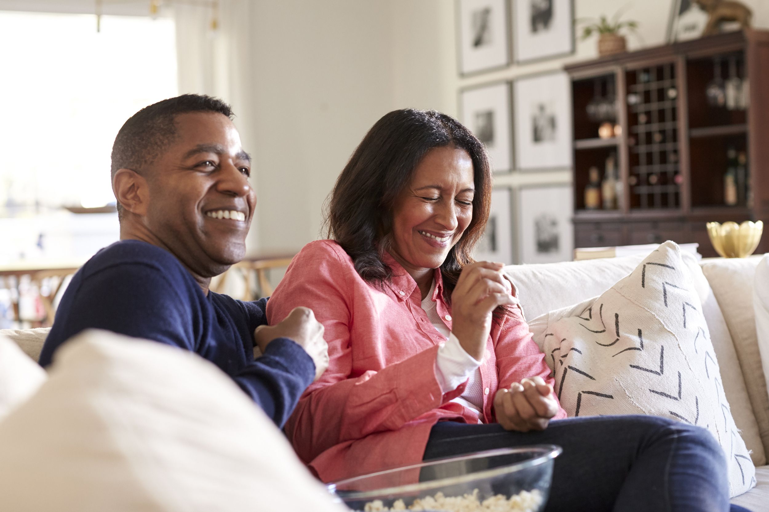 close up of middle aged couple sitting on the sofa in their living room watching TV, laughing and eating popcorn