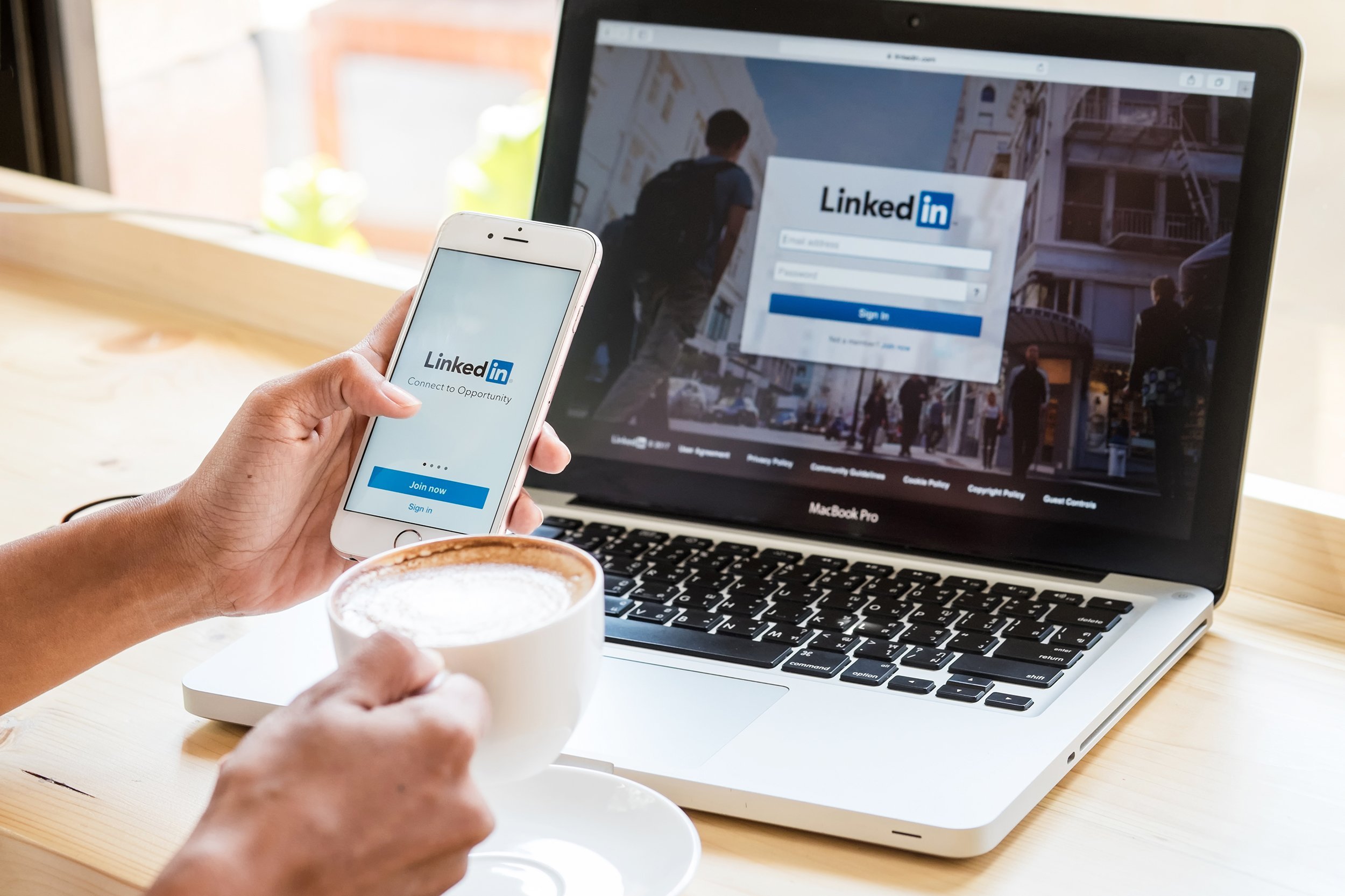 women holds Apple iPhone 6S with LinkedIn application on the screen and on laptop at coffee shop