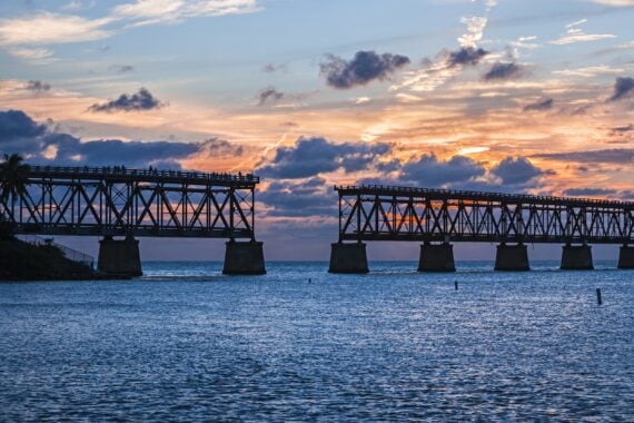 Broken steel bridge stretches over calm water at sunset, with clouds in the sky and a gap in the bridge’s span.
