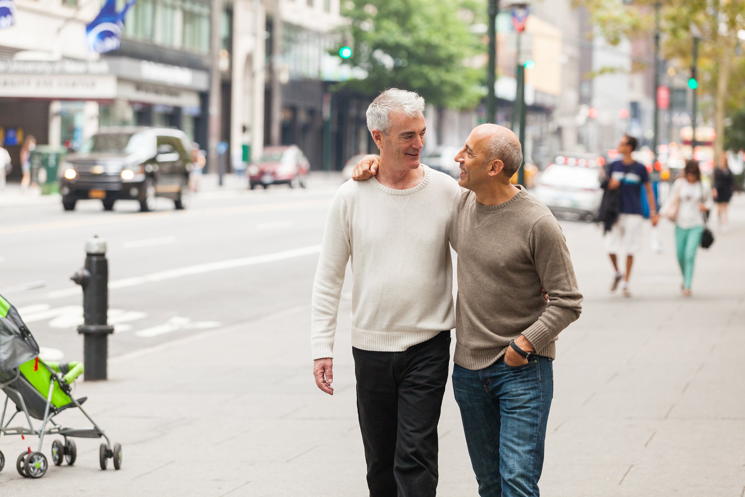 Two men walk arm in arm, smiling at each other on a city sidewalk. A green stroller and a few people are visible in the background, along with cars and tall buildings.