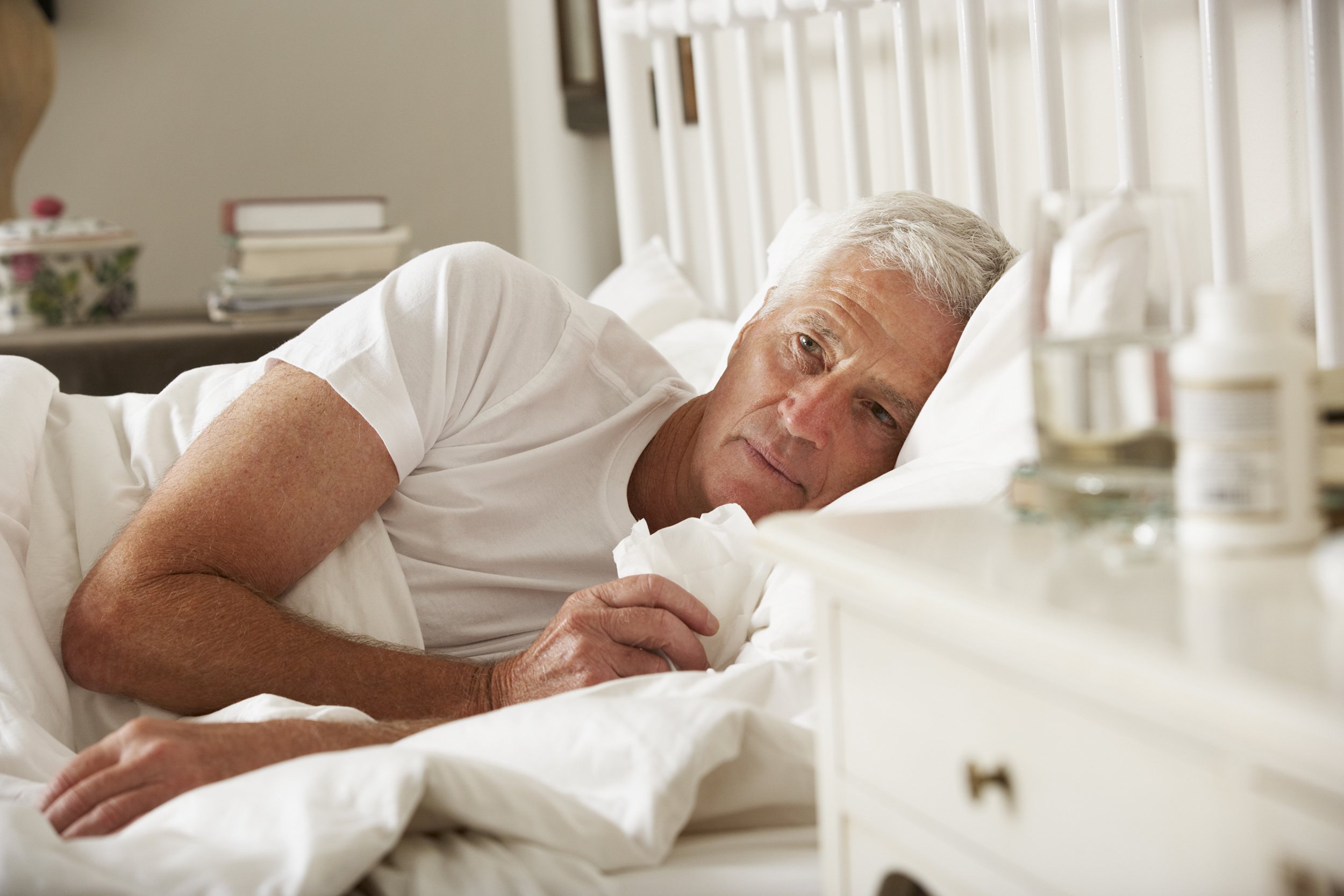 An older man with gray hair lies in bed, looking tired and holding a tissue. A bedside table with medicine bottles, a glass of water, and other items is in the foreground.