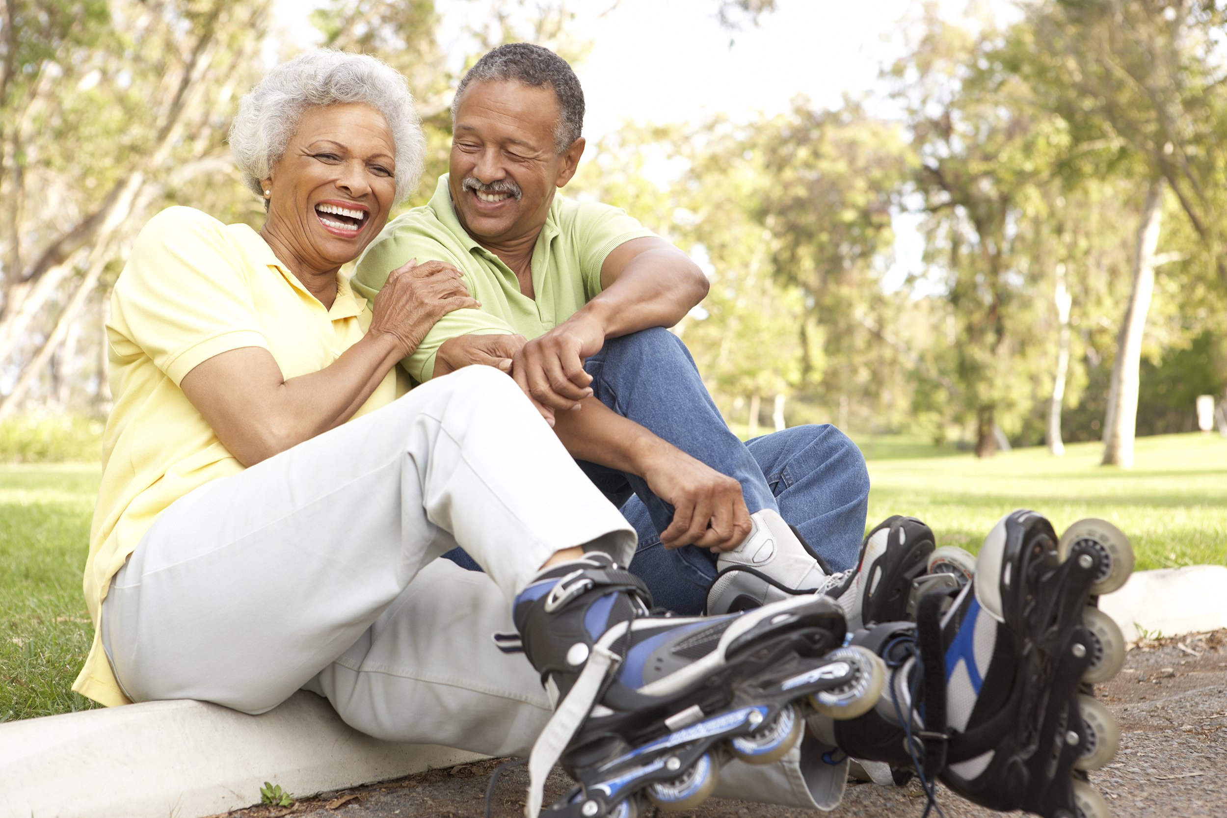 An older couple sits on the grass in a park, smiling and laughing while wearing rollerblades. They lean close to each other, enjoying a sunny day outdoors surrounded by trees.