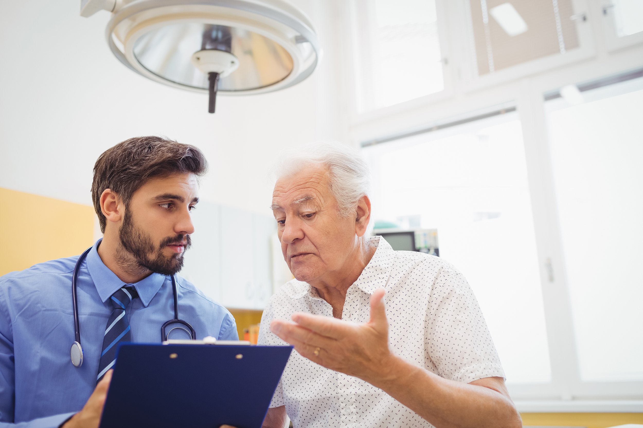 A doctor with a stethoscope around his neck discusses medical information on a clipboard with an older male patient in a well-lit examination room.