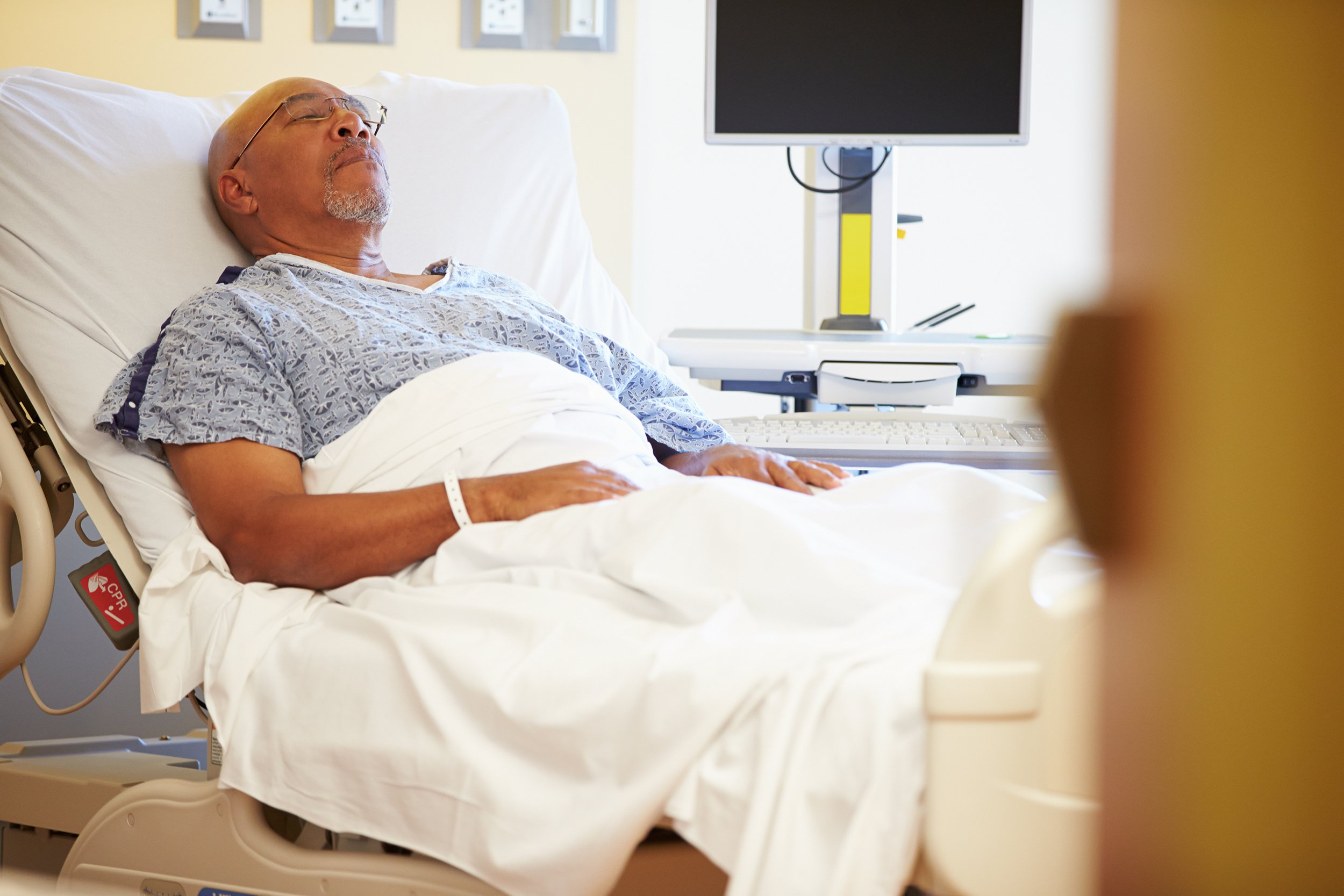 An older man wearing a hospital gown lies in a hospital bed with his eyes closed, possibly resting or asleep. Medical equipment and a monitor are visible in the background.
