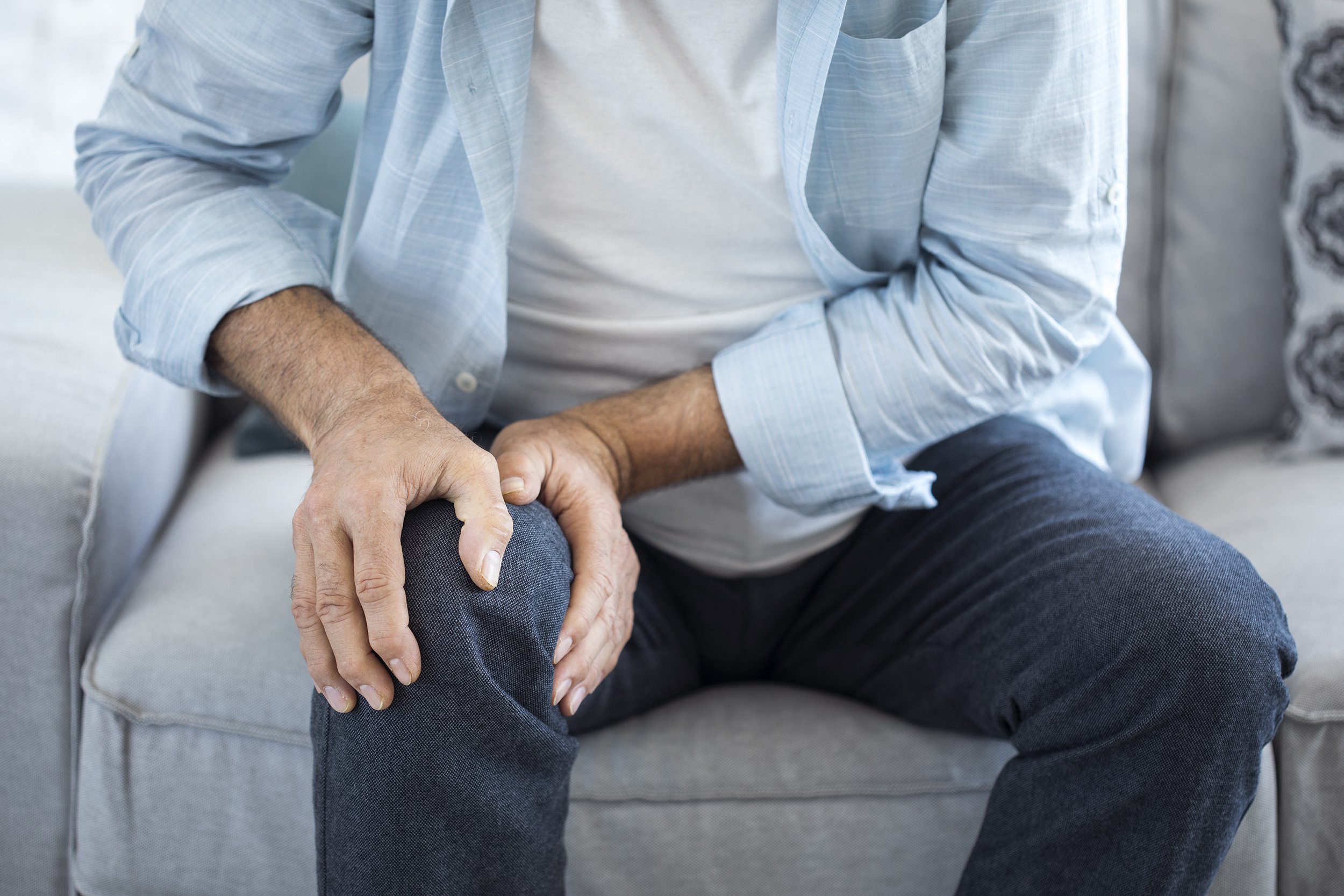 A person sitting on a couch holds their knee with both hands, appearing to experience knee pain or discomfort. The individual is wearing a light blue shirt and dark pants.