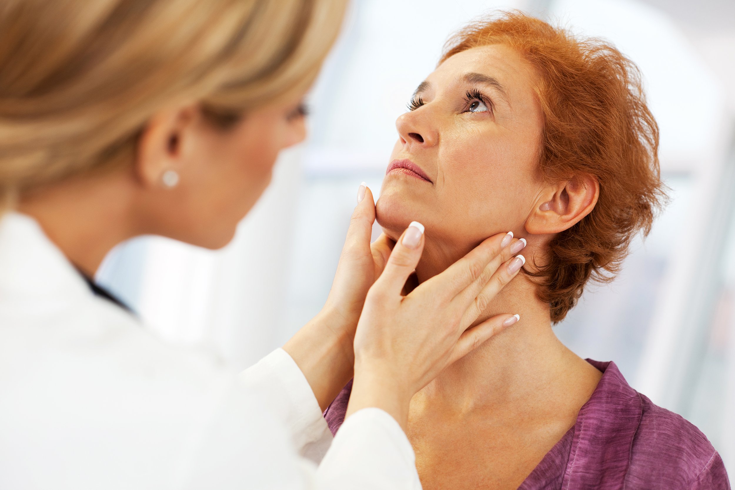 A healthcare professional examines a middle-aged woman’s neck by gently touching her throat, possibly checking her lymph nodes or thyroid. The woman looks slightly upward while being evaluated in a clinical setting.
