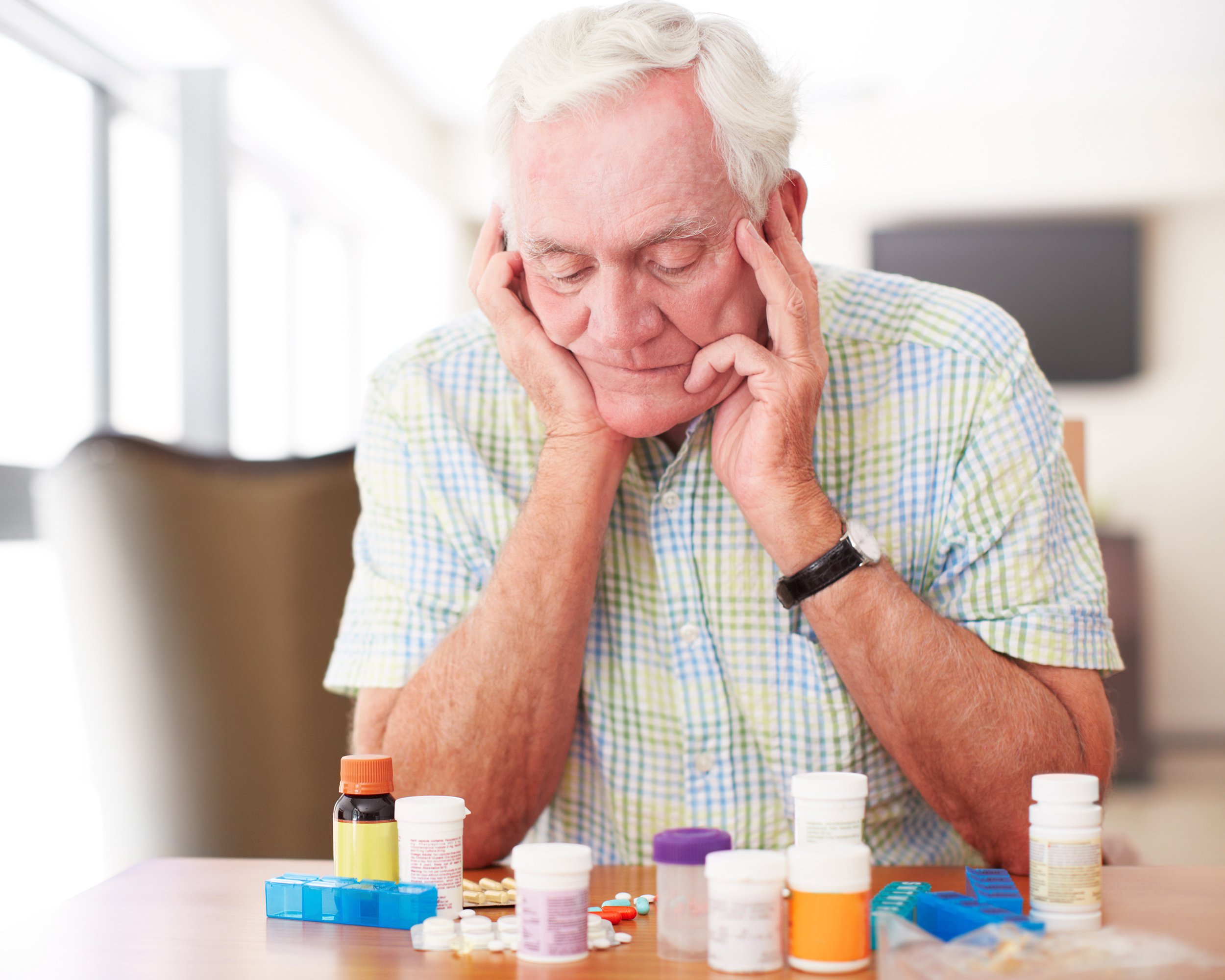 An elderly man sits at a table with his hands on his temples, looking down at several pill bottles and medication organizers, appearing thoughtful or concerned.