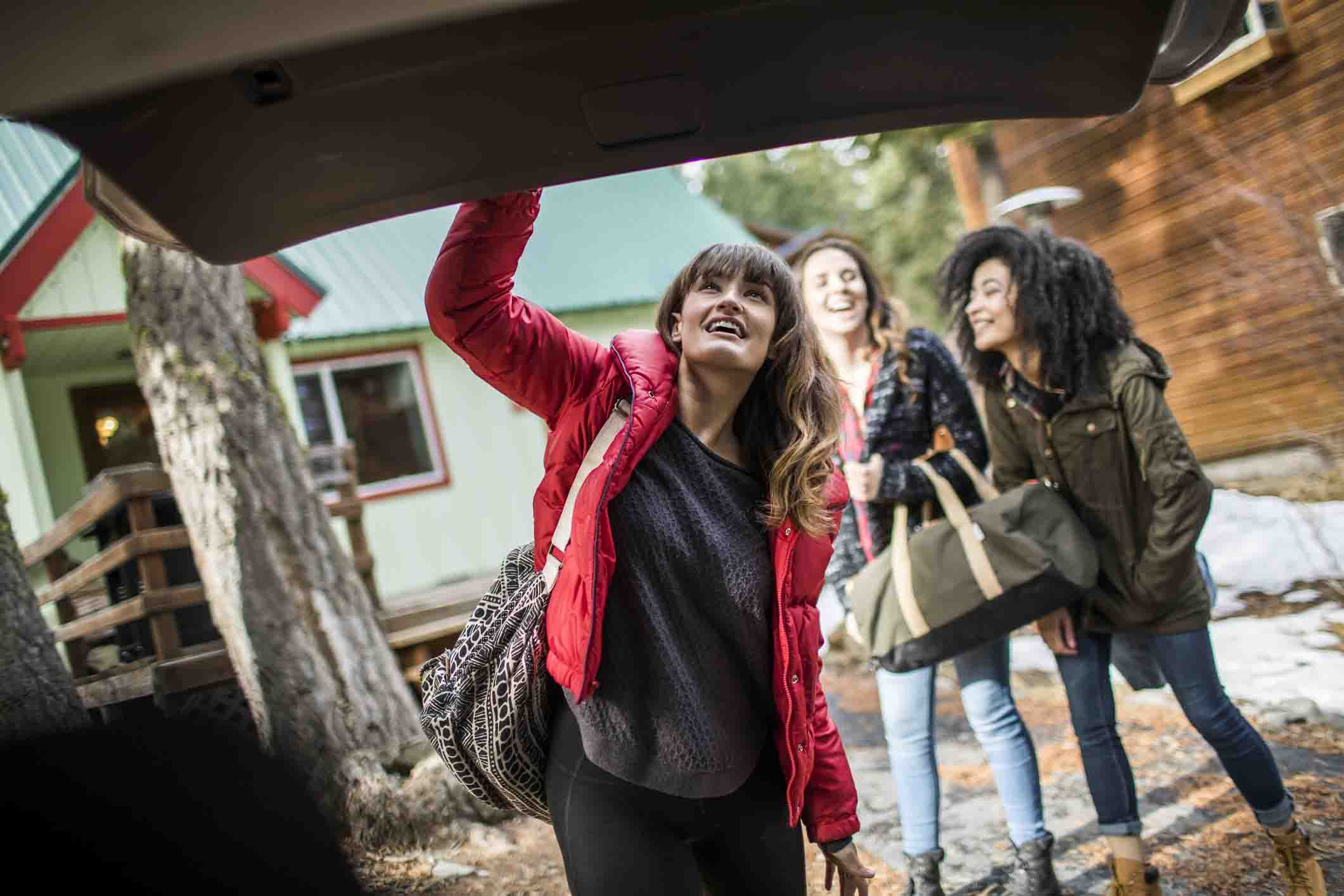 young woman shutting car trunk; friends in background at winter cabin