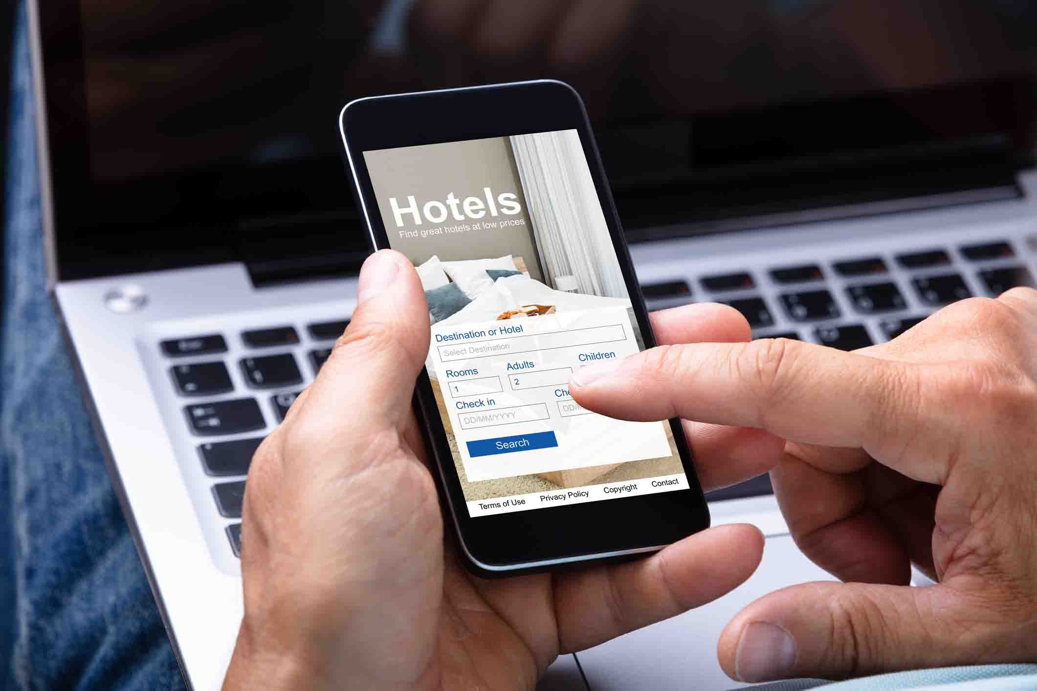 hand holding smartphone with hotel reservation app open; laptop in background