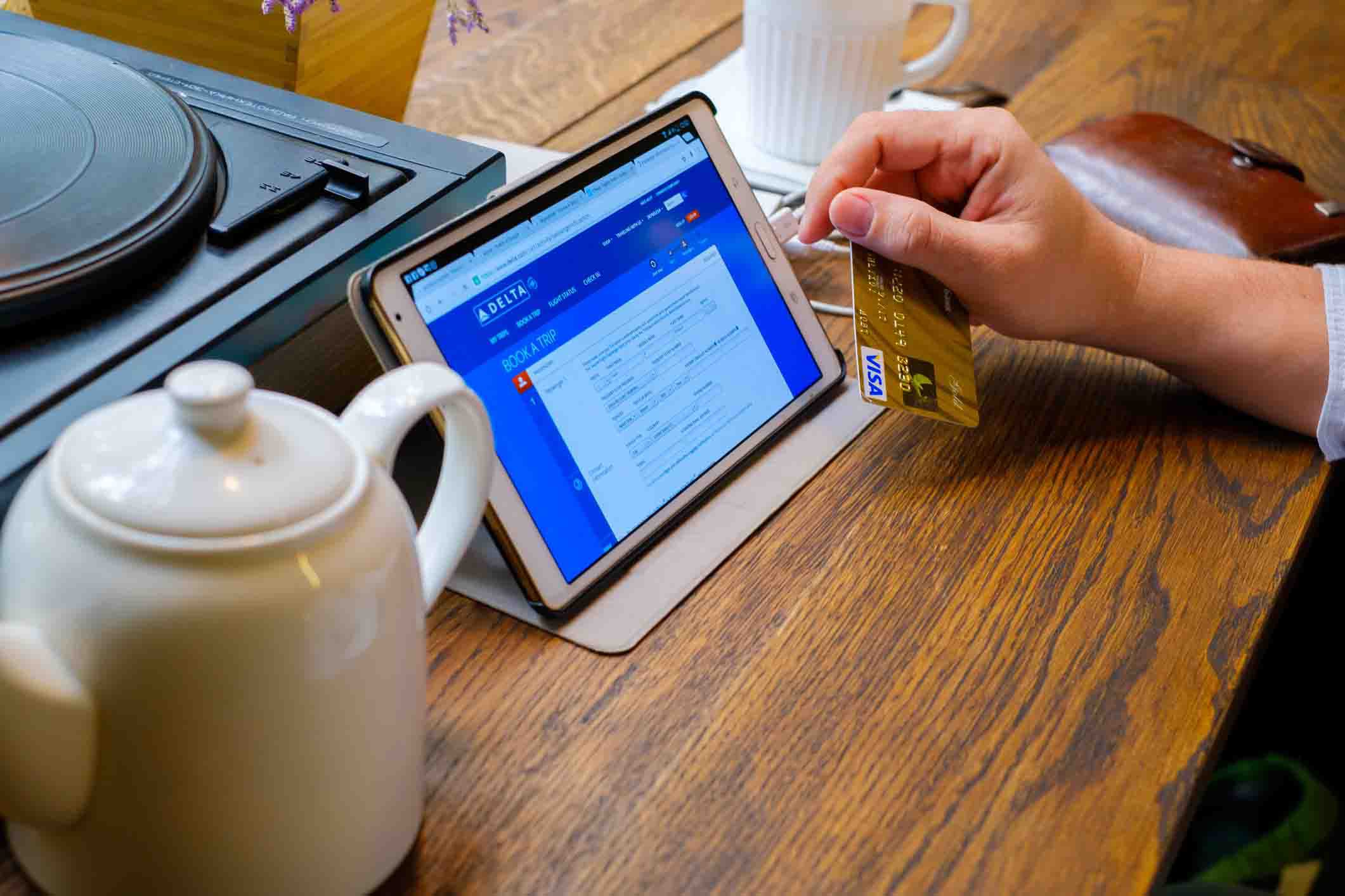 hand holding credit card, tablet with delta airlines booking page open, tea pot on table beside it