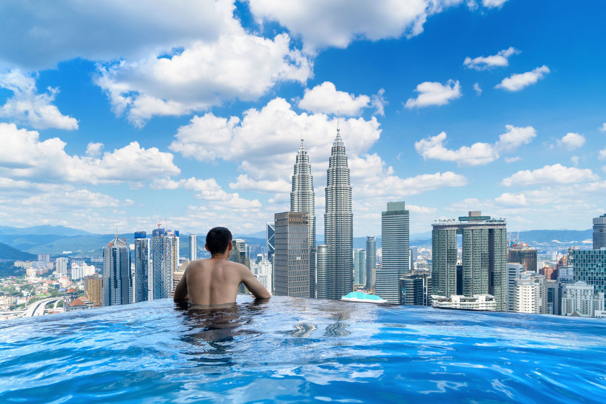 back of tourist in a swimming pool on rooftop with kuala lumpur downtown view and blue sky