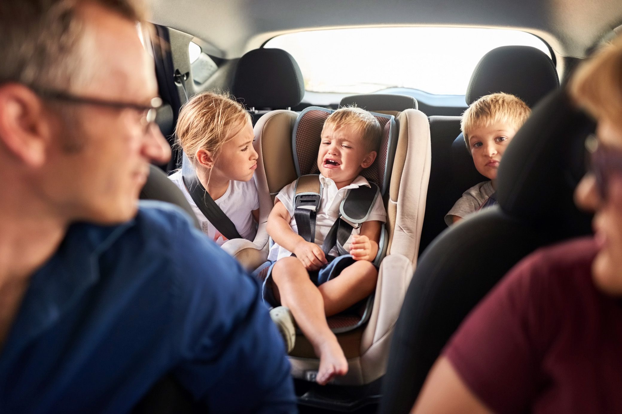 toddler crying in car with two other children and two adults trying to comfort him